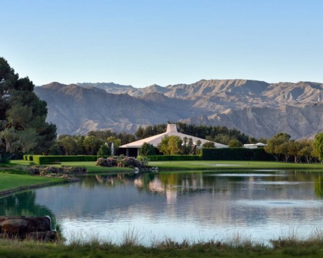 A tranquil golf course with a small pond reflecting the surrounding trees and a clear view of distant mountains against a blue sky.