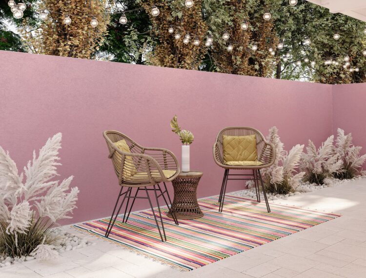 A cozy outdoor seating area featuring two woven chairs with yellow cushions on a striped multicolored rug, a small decorative table, potted plants, and white foliage. The space is enclosed by a pink wall and is adorned with hanging string lights above, set against a backdrop of greenery.
