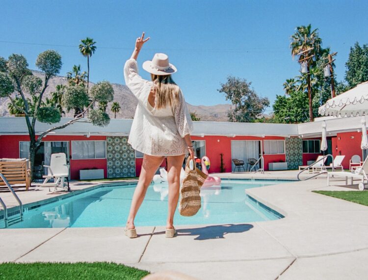 A woman in a white dress and sunhat is standing near a swimming pool with her back to the camera, making a peace sign gesture with one hand and holding a straw hat with the other. The pool area is surrounded by lounge chairs, umbrellas, and has a backdrop of red motel-style buildings with mountains and palm trees in the distance.