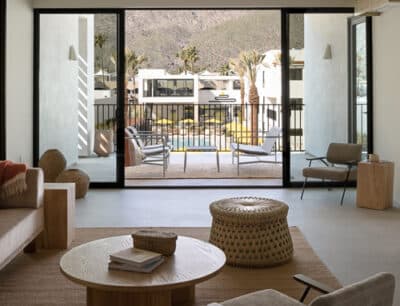 A modern living room with natural light, featuring a round wooden coffee table, woven chairs, and a sofa. Sliding glass doors open to an outdoor patio with furniture overlooking a scenic mountain landscape.