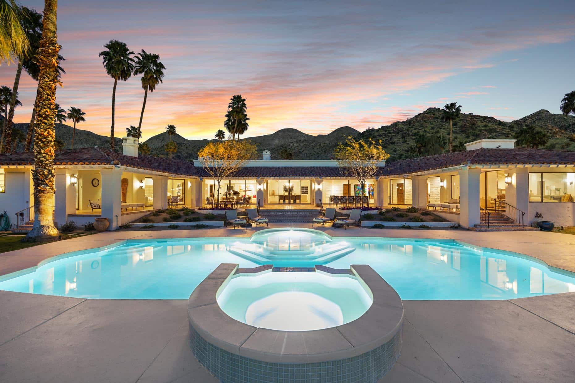 A luxurious pool area in front of a single-story villa at twilight, with lit interior rooms, outdoor furniture, palm trees, and a mountainous backdrop under a colorful sunset sky.