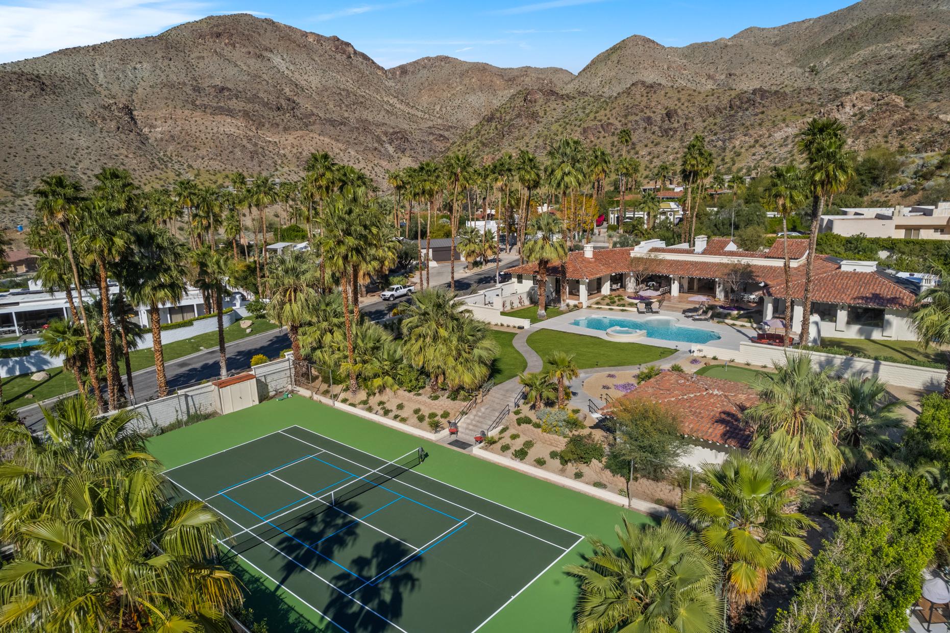 An aerial view of a luxurious resort with a tennis court in the foreground, a swimming pool surrounded by palm trees, and a mountainous backdrop.