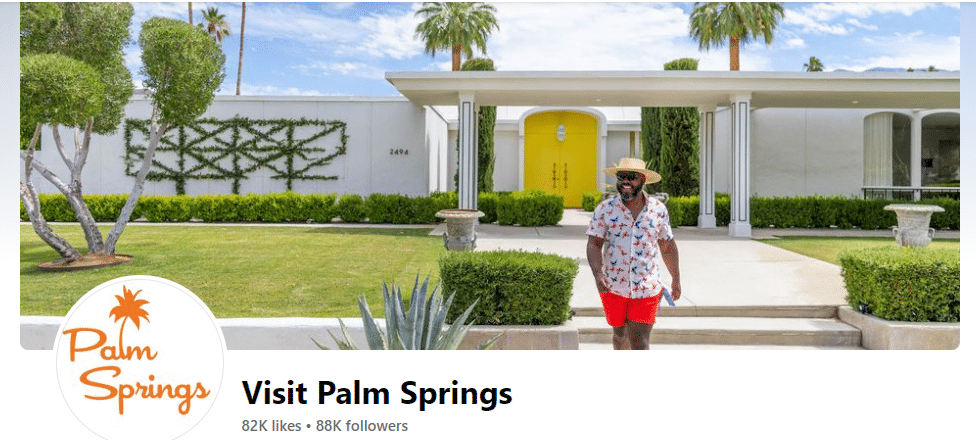 A person in a patterned shirt and shorts walking in front of a white mid-century modern style building with a yellow door, under a clear blue sky in Palm Springs, as indicated by the "Visit Palm Springs" overlay with a palm tree logo in the lower left corner.