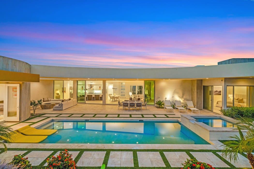 A luxurious backyard at dusk featuring a symmetric rectangular swimming pool surrounded by neatly trimmed grass and flower beds, with a modern single-level house with large windows and sliding doors, outdoor furniture, and a vibrant pink and blue sky in the background.