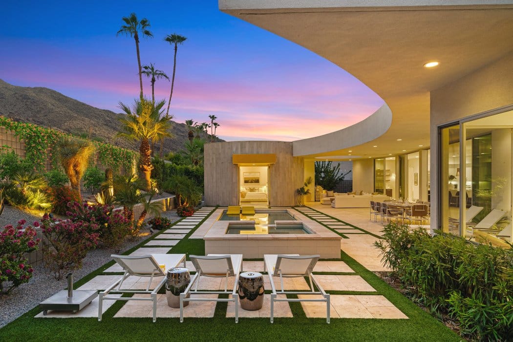 A modern outdoor patio with seating and a fire pit, overlooking a geometric garden and situated against a backdrop of a pink and blue sky at dusk, with palm trees and a mountainous landscape in the distance.