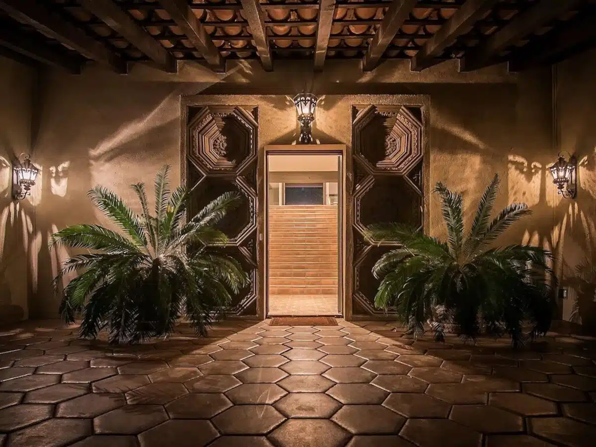 Elegant entrance of a building featuring an ornate wooden door set in a textured wall with symmetrical geometric designs, flanked by ornamental sconces and lush potted palms, under an exposed wooden beam ceiling with terracotta tiled flooring.