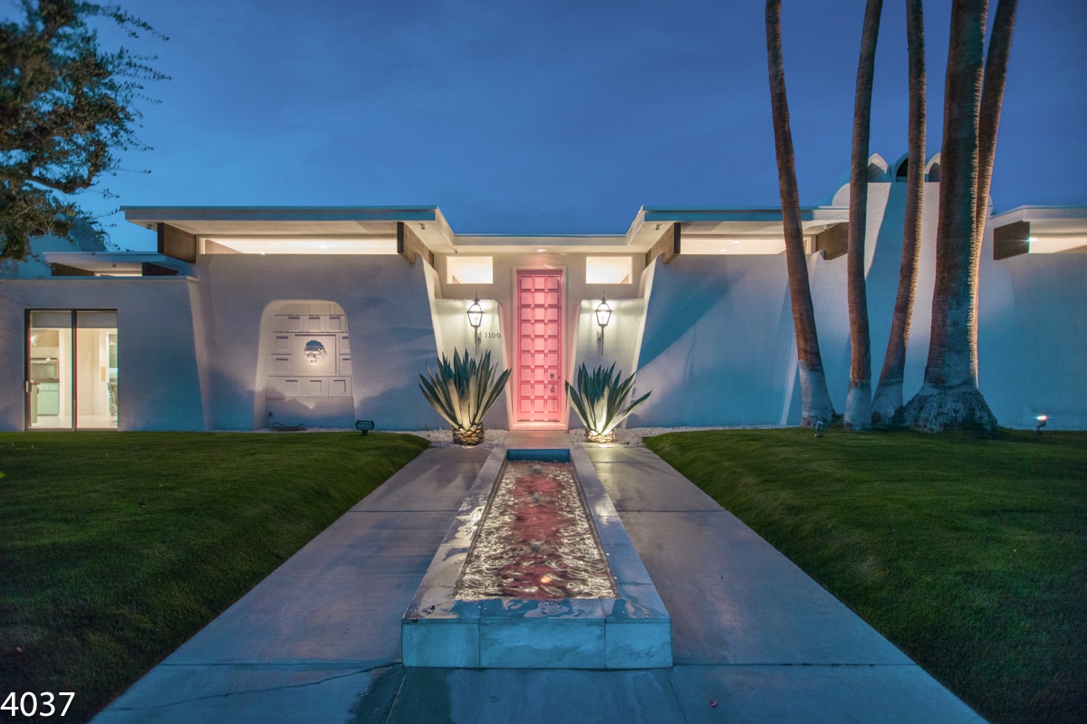 A modern house exterior at dusk with a central pink door lit by lanterns, flanked by two large agave plants and a pathway with a water feature, with tall palm trees to the right and a neat lawn around.