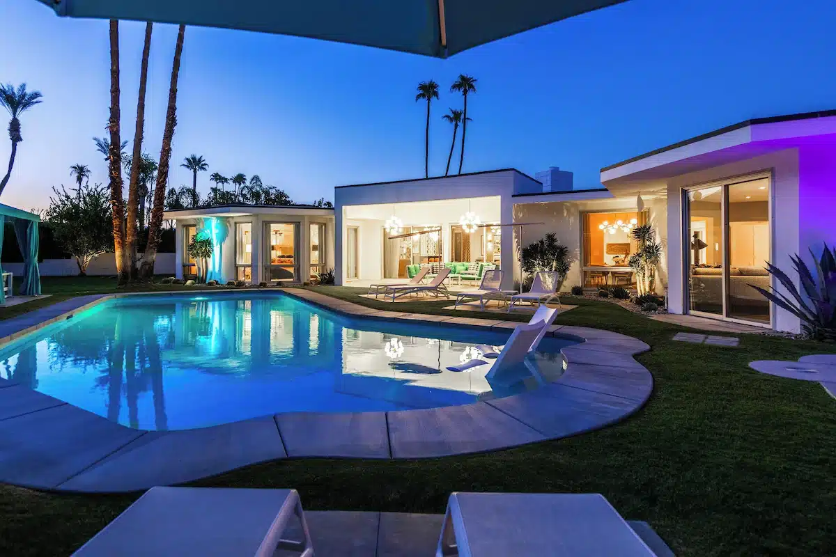 A modern house with illuminated interiors and exterior LED lights at twilight, featuring a swimming pool, lounge chairs, and palm trees in the background.