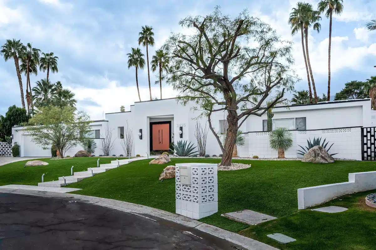 A white mid-century modern style house with a red front door, surrounded by palm trees, a manicured lawn with decorative stones and desert plants, under a cloudy sky.