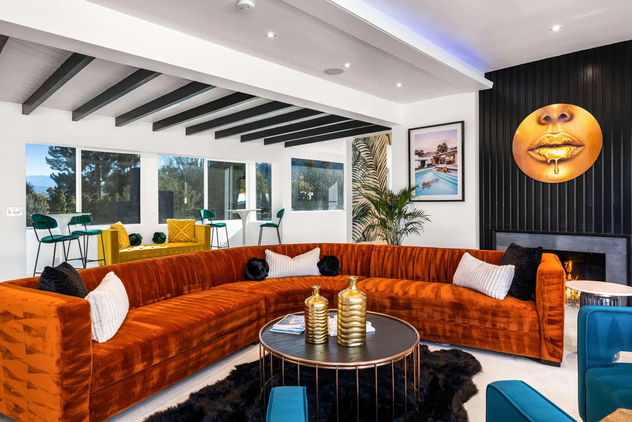 A modern living room with a large orange velvet sectional sofa, contrasting black and white throw pillows, a circular coffee table with golden vases, and a fireplace. The décor includes a dark paneled wall with a round golden artwork depicting lips, colorful chairs at a bar area by the window, and vibrant plants and framed artwork. The room features white walls with exposed gray beams on the ceiling and ample natural light.