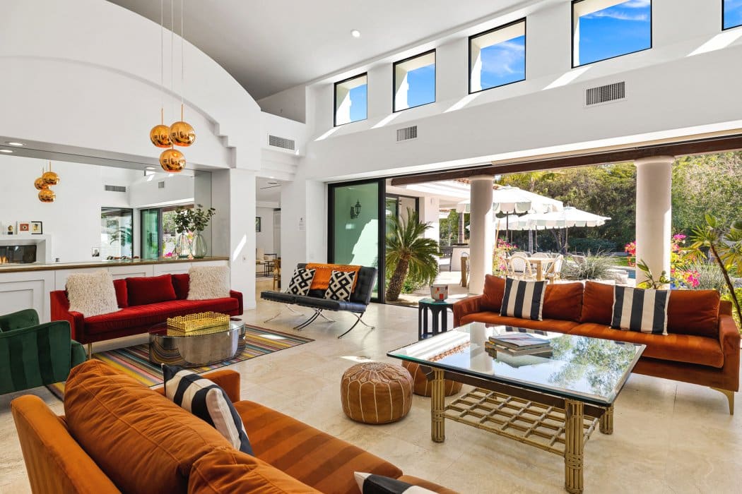Bright and spacious modern living room with high ceilings, large windows, and an open layout that leads to an outdoor patio area with umbrellas and seating. The room features multiple colorful sofas, decorative lighting, and a mix of eclectic furnishings.