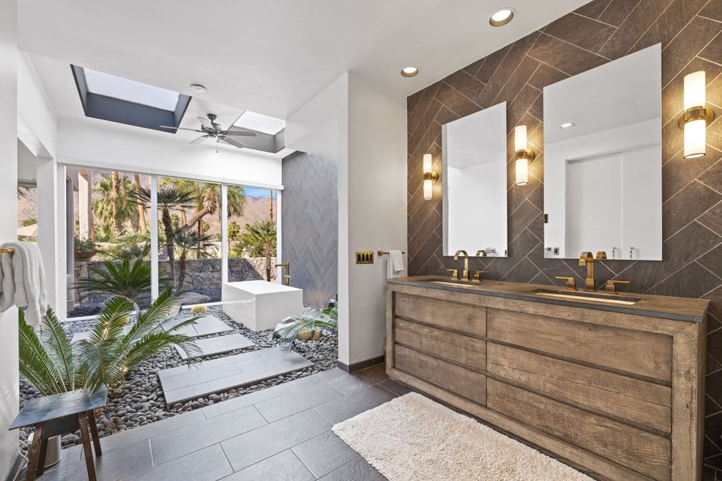 A modern bathroom featuring a freestanding tub, a large walk-in shower with a glass front, double sinks with gold fixtures on a wooden vanity, two wall-mounted mirrors, herringbone tile walls, and a floor-to-ceiling window with a view of palm trees and mountains. A ceiling fan, skylight, and stylish wall sconces provide ample lighting. Decorative pebbles, stepping stones, and greenery create a natural ambiance in the shower area.