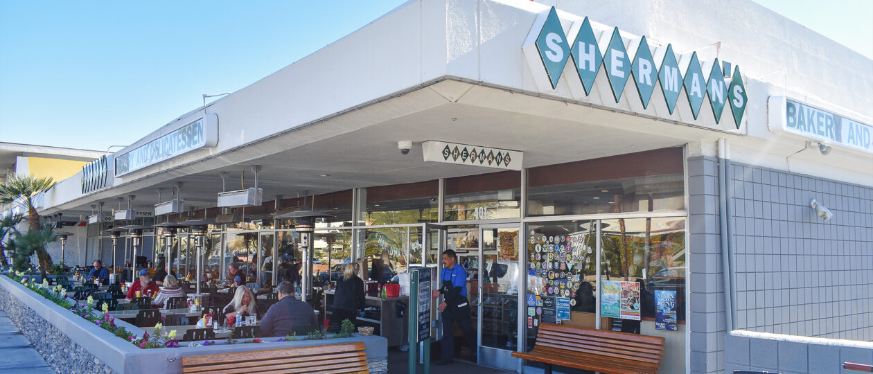 Alt text: Exterior view of Sherman's Deli and Bakery with patrons dining outside on a sunny day. A variety of stickers are displayed on the window, and there's a clear blue sky overhead.