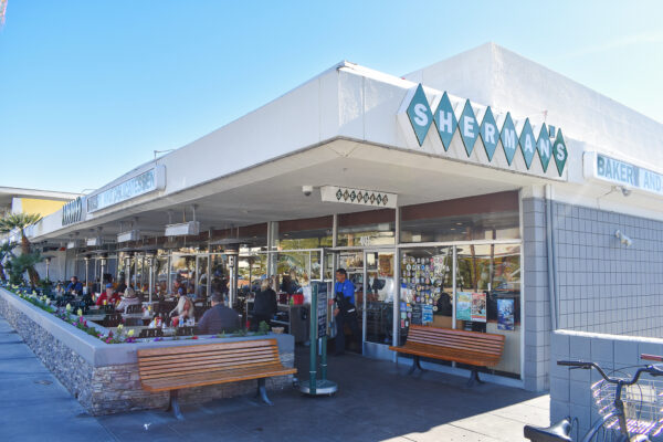 Alt text: Exterior view of Sherman's Deli and Bakery with patrons dining outside on a sunny day. A variety of stickers are displayed on the window, and there's a clear blue sky overhead.