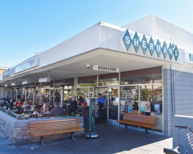 Alt text: Exterior view of Sherman's Deli and Bakery with patrons dining outside on a sunny day. A variety of stickers are displayed on the window, and there's a clear blue sky overhead.